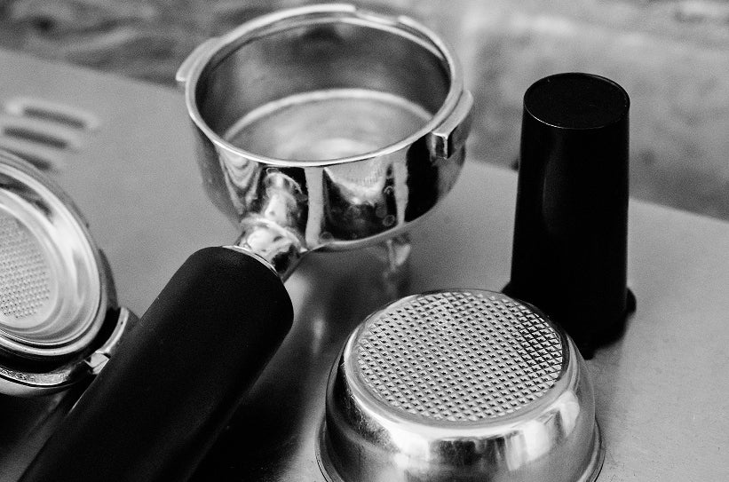 Multiple portafilter baskets and tamper laid out on a surface in black and white