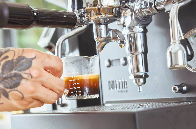 A tattooed hand holding a graduated shot glass under a commercial espresso machine group head