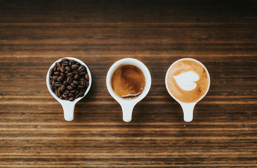 Three white cups in a row showing whole coffee beans, a straight espresso shot, and a latte with latte art