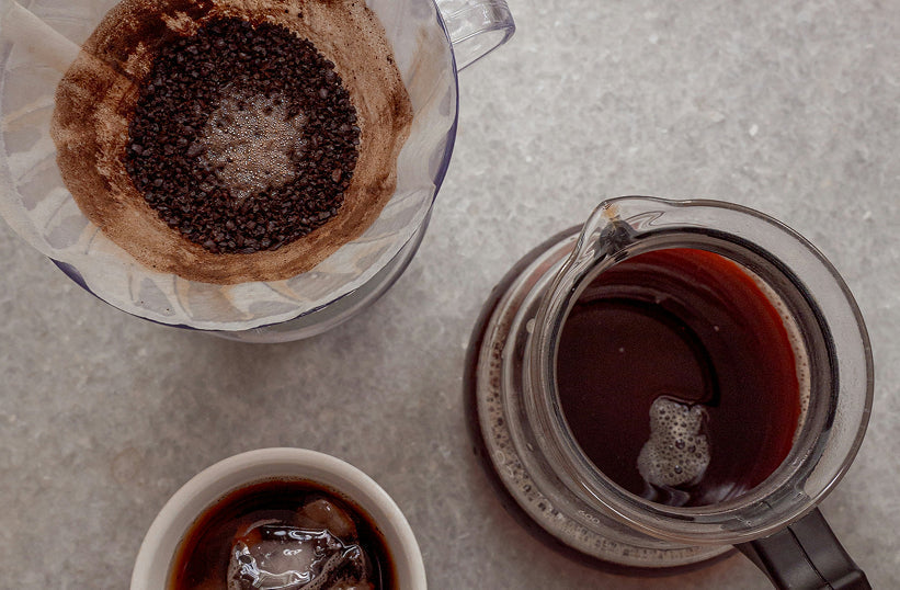An overhead view of a pour over dripper with used coffee grounds next to a glass server and espresso cup