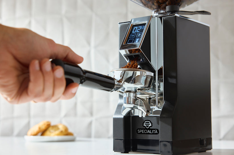 A hand holding a portafilter under a Eureka Specialità espresso grinder dispensing fresh grounds