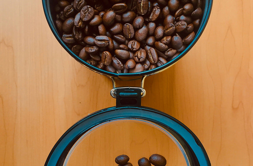 An open airtight canister filled with whole coffee beans, lid propped open on a wood surface