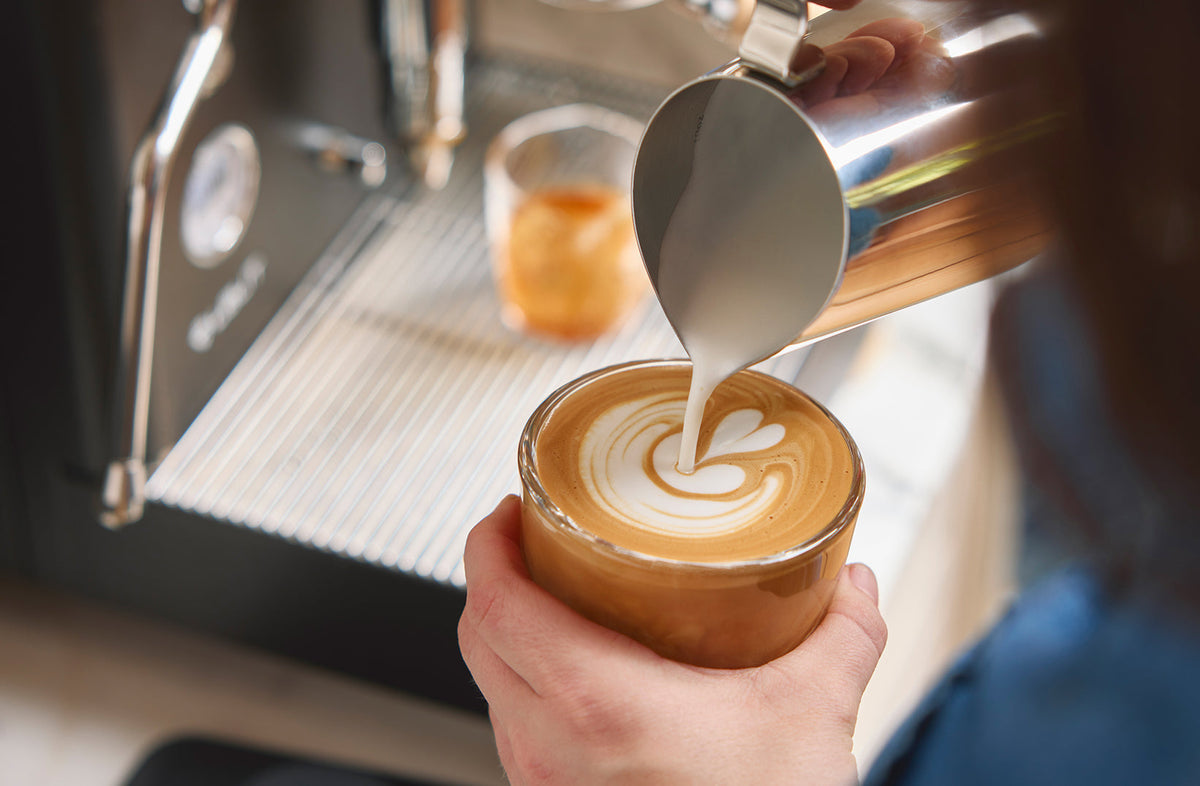 Barista pouring steamed milk with latte art into a glass espresso cup at a home machine - Seattle Coffee Gear