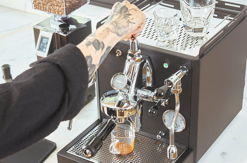A tattooed hand operating the lever on an E61 group head espresso machine as espresso extracts into a shot glass