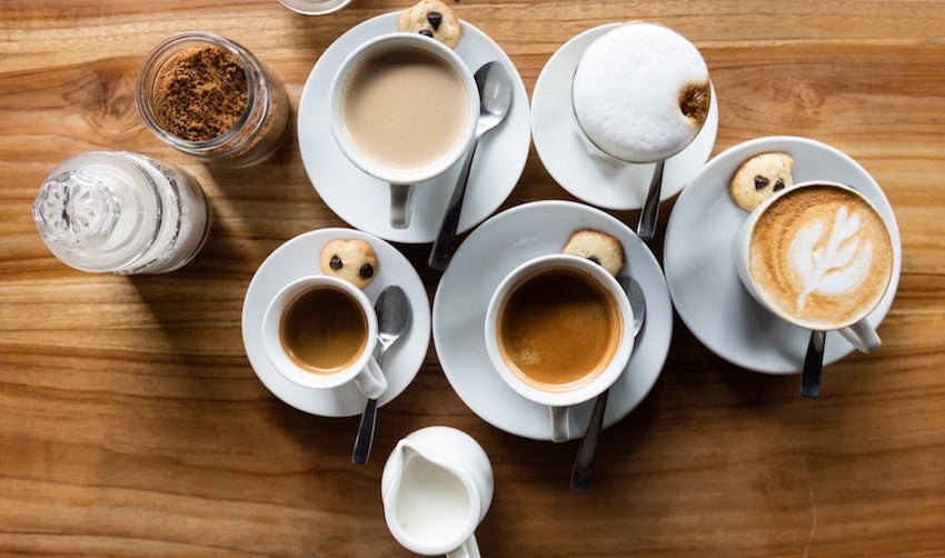A flat lay of coffee drinks including lattes and espresso alongside a sugar bowl, milk jug, and spoons arranged on a table