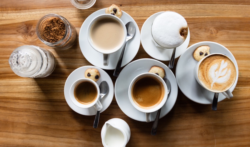A flat lay of coffee drinks including lattes and espresso alongside a sugar bowl, milk jug, and spoons arranged on a table