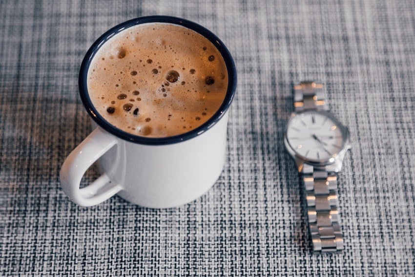 A cup of coffee topped with milk foam on a checkerboard plaid tablecloth, with a wristwatch resting beside it